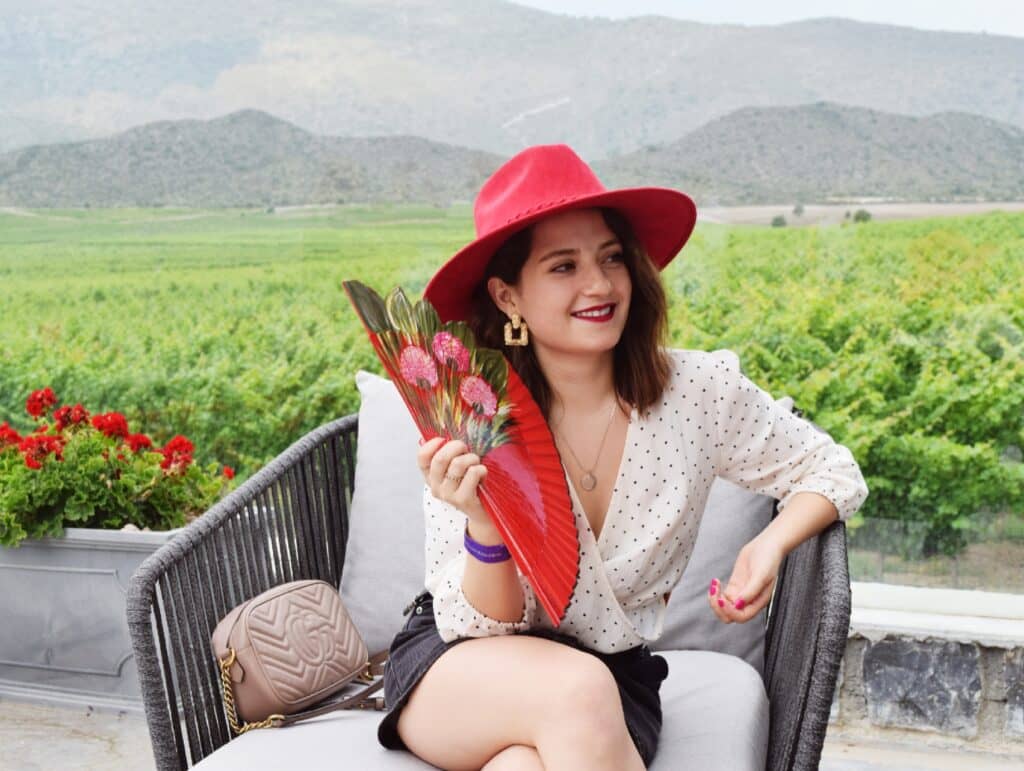 mujer con sombrero rojo y abanico rojo en terraza con montañas detrás 