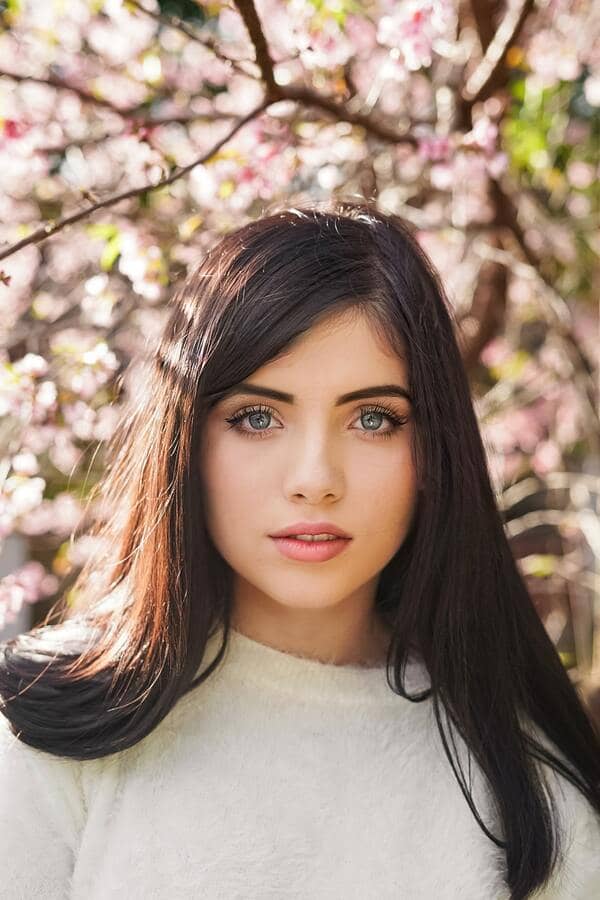 Mujer con cabello largo oscuro y ojos azules, con un árbol de flores rosas detrás de ella