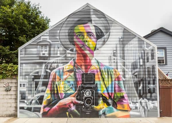 Arte urbano de un hombre sosteniendo una cámara, pintado en la fachada de una casa.