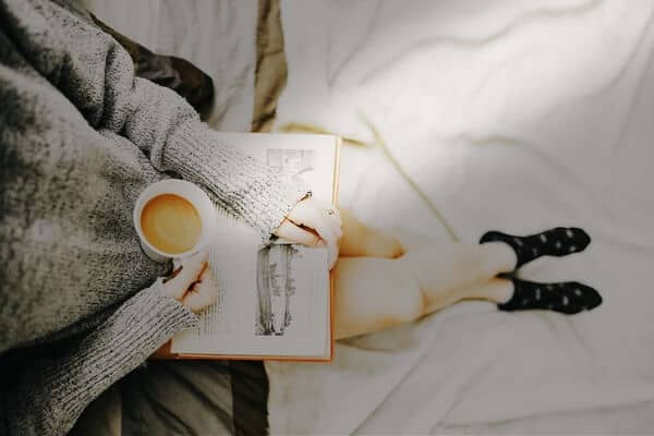 Foto tomada desde arriba de una persona con un libro apoyado sobre sus piernas y una taza de café.