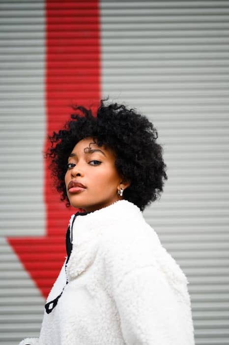 woman wearing a white jacket stands in front of white wall with a red arrow