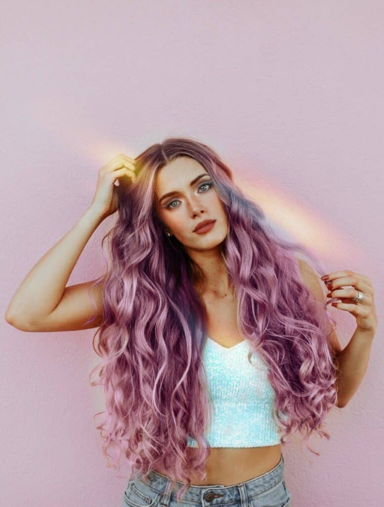 Pride edit of woman with purple hair in a white top stands in front of a pink wall