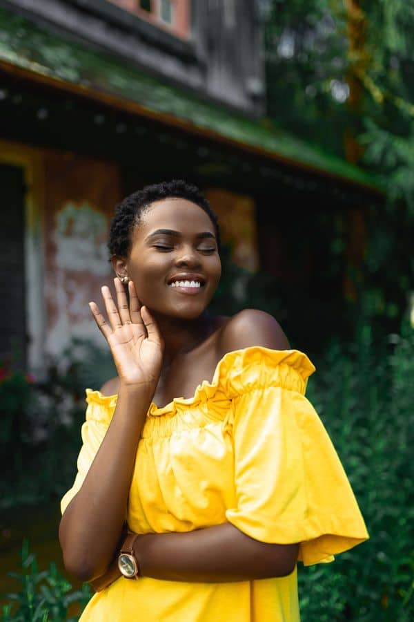 smiling woman in yellow dress stands in front of background with green foliage