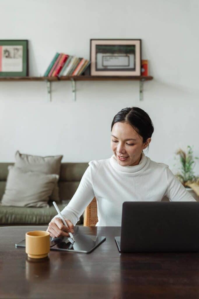 Mulher sentada, com o laptop aberto e escrevendo algo no tablet